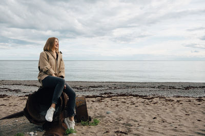 Cheerful young woman sitting on weathered metal at beach against sky