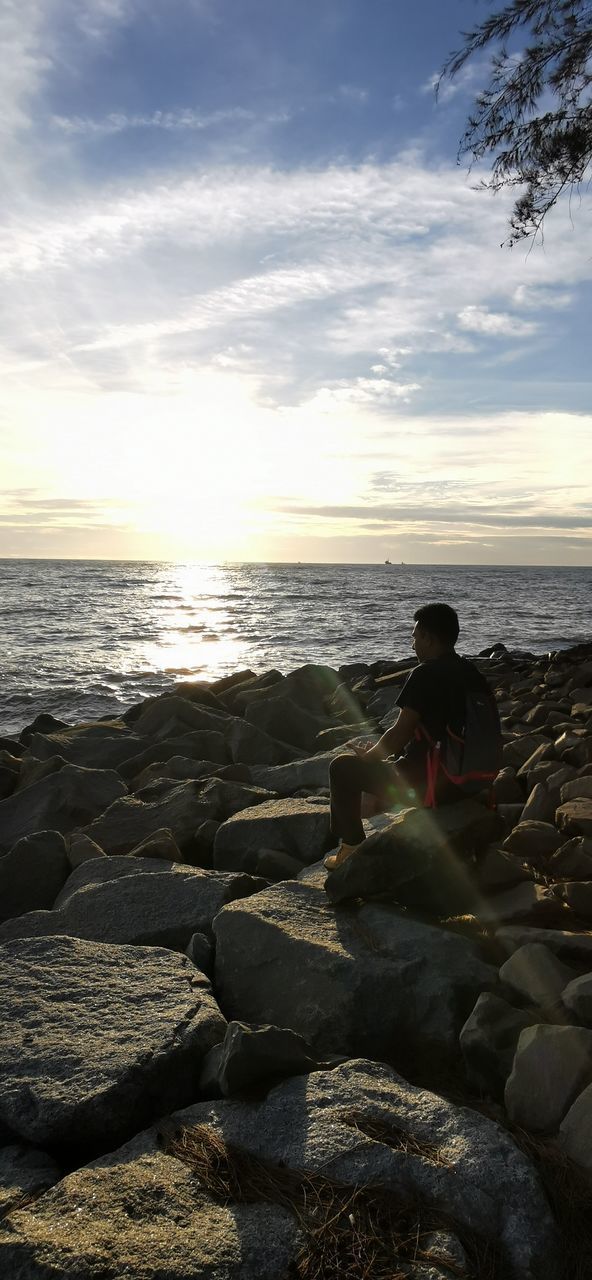 MAN SITTING ON ROCK BY SEA AGAINST SKY DURING SUNSET
