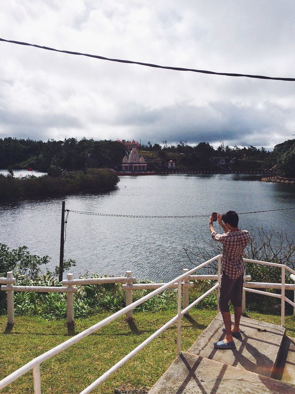 water, sky, railing, river, lifestyles, standing, leisure activity, rear view, full length, cloud - sky, lake, casual clothing, built structure, nature, architecture, cloud, building exterior, day