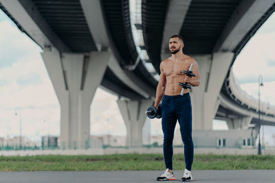 Full length of shirtless man exercising while standing under bridge