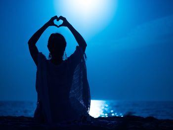 Rear view of woman making heart shape while sitting at beach during night