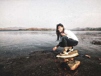 Happy young woman crouching on rock at beach against sky