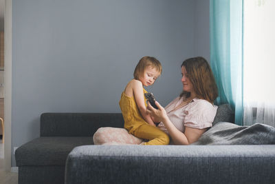 Plus, european mother hugs toddler child sitting with smartphone sofa real interior of living room