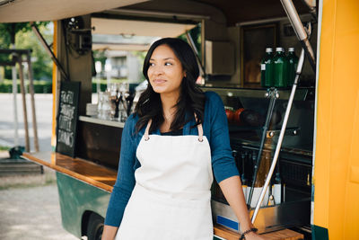 Smiling female owner looking away while standing against food truck