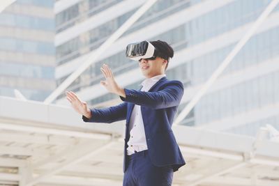 Low angle view of smiling businessman enjoying while using vr glasses against building