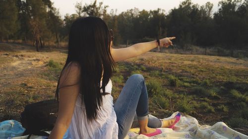 Young woman pointing while sitting on blanket against trees in park during sunset