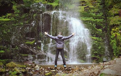 Rear view full length of man with arms outstretched standing against waterfall at forest