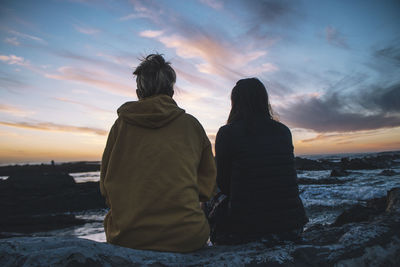 Rear view of female friends sitting on rocky shore during sunset