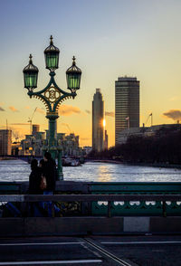 People sitting by river in city against sky during sunset