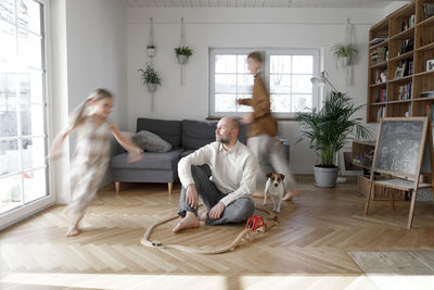 Siblings running around father sitting on floor in living room at home