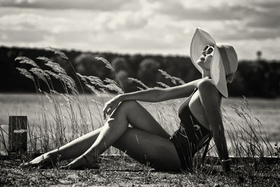 A woman in a swimsuit, hat and sunglasses sunbathes in summer on the riverbank among the grass