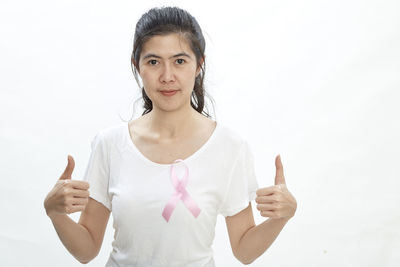 Portrait of woman showing thumbs up while wearing pink ribbon on top against white background
