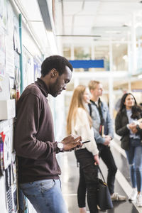 Side view of young man using mobile phone while leaning on wall at university