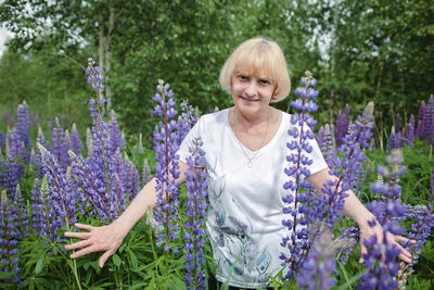 Happy attractive senior woman enjoys walk in purple lupines in blooming field, active in retirement