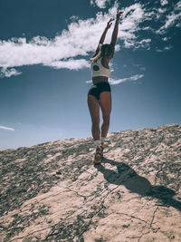Full length of young woman with arms raised standing on mountain against sky during sunny day