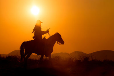 Silhouette people riding horse on field against orange sky