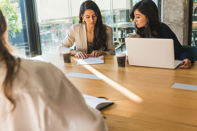 High angle of cheerful hispanic female entrepreneurs in smart casual clothes smiling and discussing business ideas while sitting at table and working on project in sunlit office together