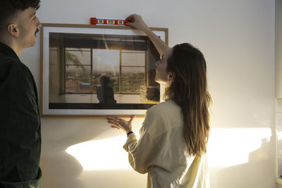 Young couple measuring wall while hanging picture frame in new house