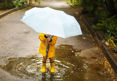 Low section of boy holding umbrella standing in puddle during rainy season