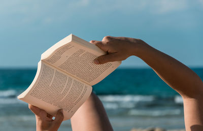 Low angle view of hand holding a book at beach against sky