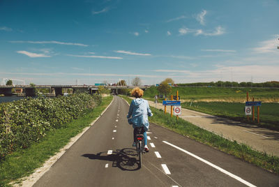 Rear view of woman riding bicycle on road amidst plants against sky