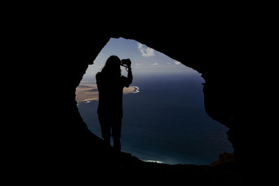 Silhouette man photographing in cave against sky