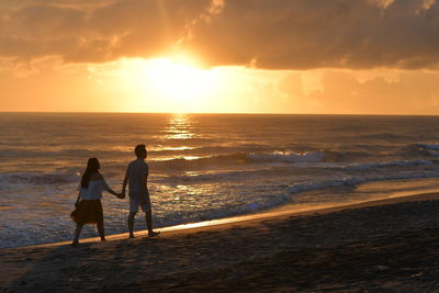 Rear view of friends walking on beach against sky during sunset