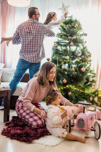 Excited father with daughter on hands decorating fir tree with star in living room with mother and toddler playing with toy and velomobile