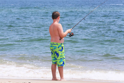 Full length of shirtless boy fishing in sea while standing on shore at beach