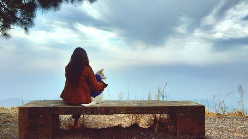 Rear view of woman sitting on a bech with dog against sky