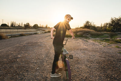 Side view of young man with bicycle standing on field against clear sky during sunset