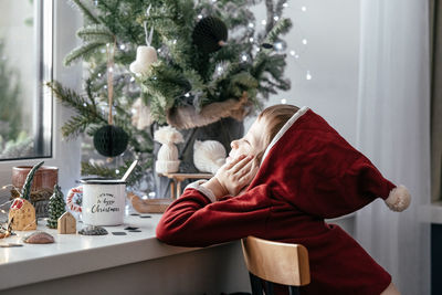 A thoughtful boy in a santa costume sits at the window and makes christmas decorations