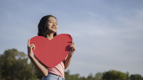 Portrait of smiling woman holding heart shape against sky