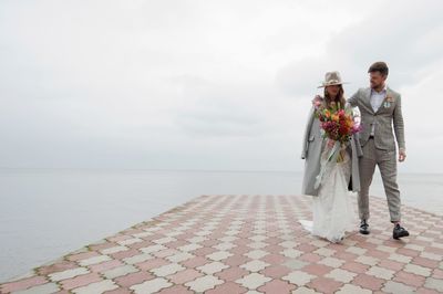 Newly married couple walking on pier over lake against cloudy sky