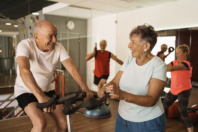 Cheerful senior woman in activewear laughing while standing near motivated husband exercising on cycling machine during workout in modern gym