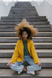 Afro woman with tousled hair meditating while sitting on staircase