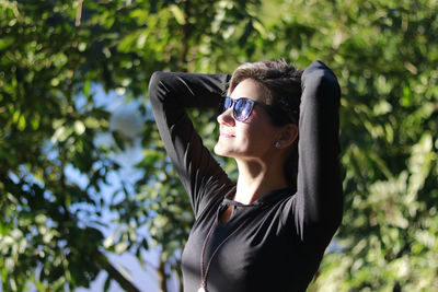 Woman with hands behind head wearing sunglasses against trees