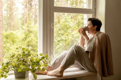 Middle age beautiful woman sitting on windowsill, drinking tea, dreaming. 50-year-old woman relaxing