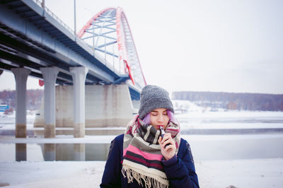 Young woman using phone while standing on snow covered bridge