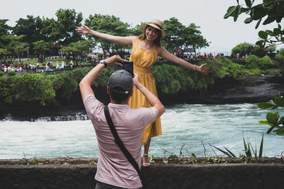 Woman with arms raised standing by lake against trees