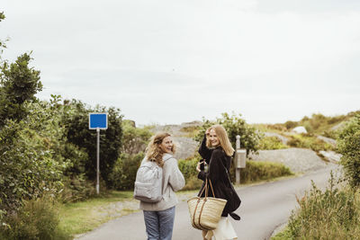 Smiling female friends looking over shoulder while walking on road during summer vacation