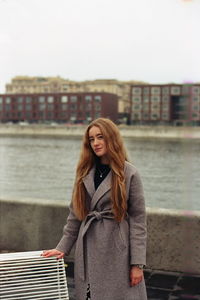 Portrait of young woman standing against river and buildings against sky