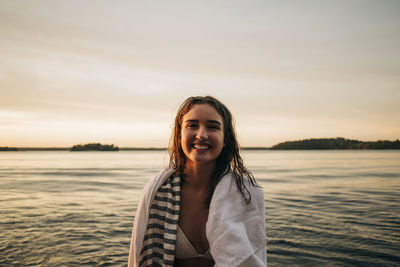 Portrait of happy woman with towel against lake during sunset