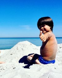 Portrait of shirtless boy showing thumbs up while sitting on beach against clear blue sky during sunny day