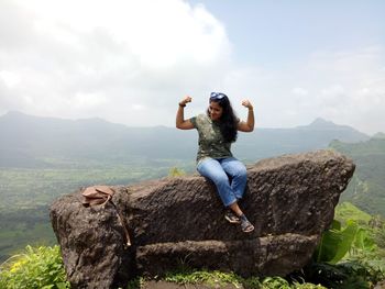 Full length of young woman sitting on rock against sky