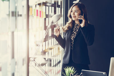 Smiling businesswoman talking over smart phone while standing by window in office