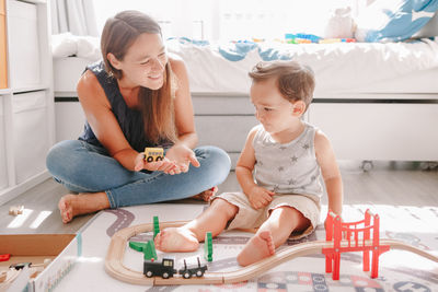 Mother and toddler boy playing with car wooden railway on floor at home. early age education 