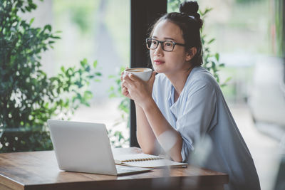 Thoughtful woman having coffee while standing by laptop at table