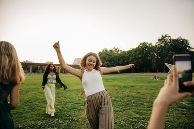 Teenage girl photographing carefree female friend dancing at park during sunset