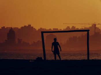 Silhouette man standing at beach by goal post against clear sky during sunset
